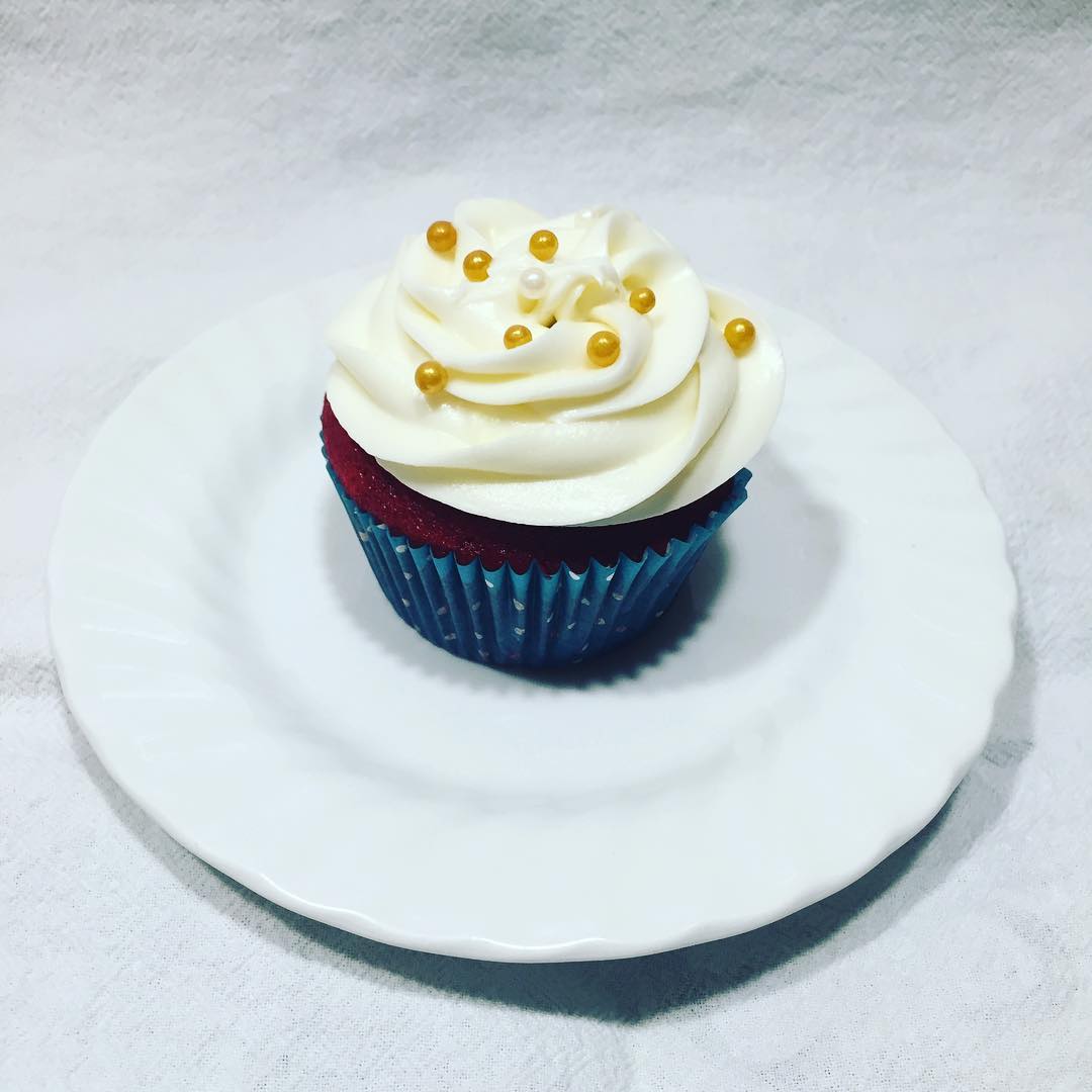 red velvet cupcake with white cream cheese icing and golden sugar pearls on a white ceramic plate with a white fabric background